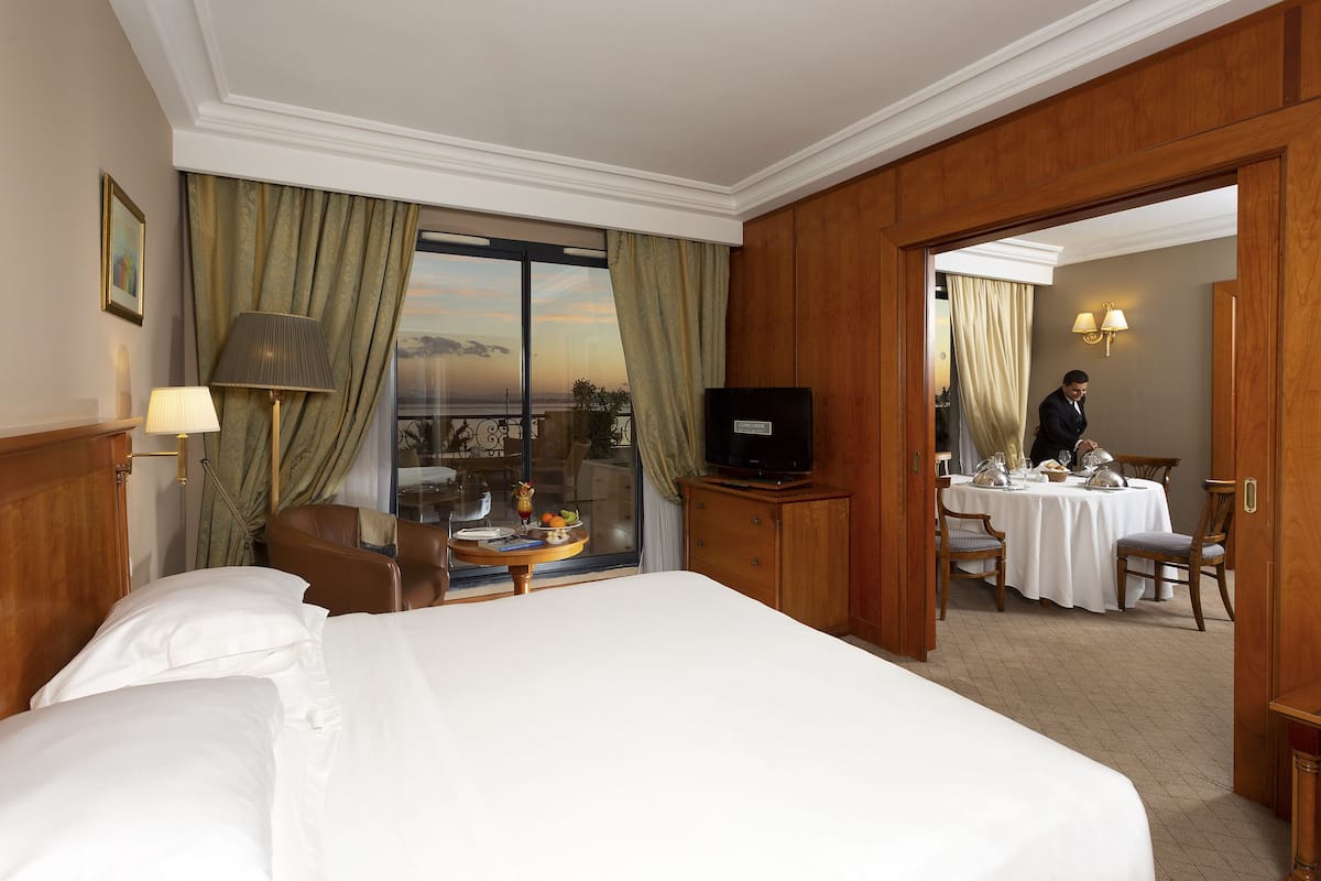 Une chambre d’hôtel avec un grand lit, une télévision, un coin repas avec une table dressée pour deux personnes, et une vue par la fenêtre.