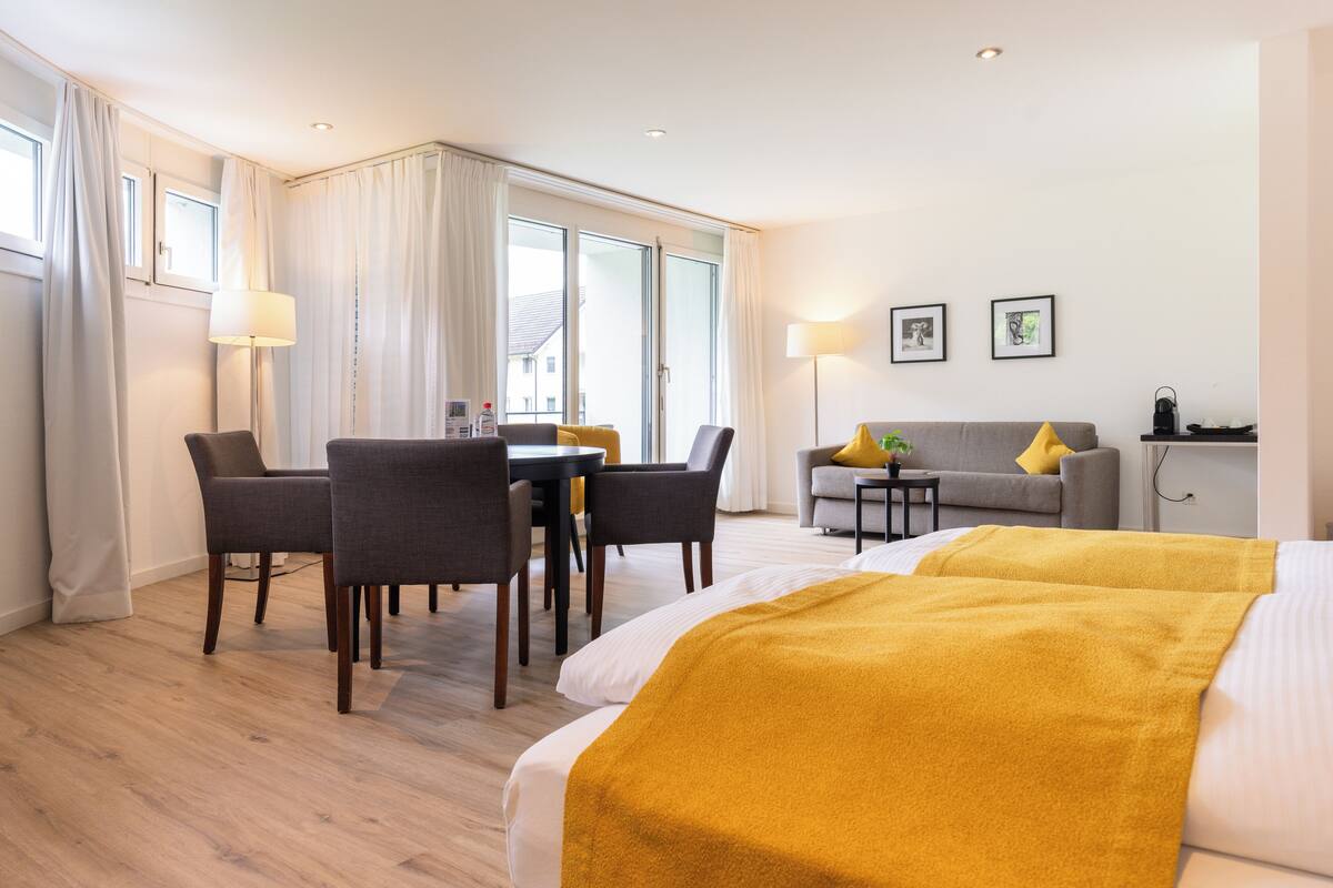 Ein Hotelzimmer mit einem Bett, einem Sofa, einem Esstisch mit Stühlen und einem Schreibtisch mit Lampe.