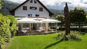 A two-story house with a balcony, outdoor seating area with umbrellas, and a garden. - Hotel Schatzmann (Triesen)