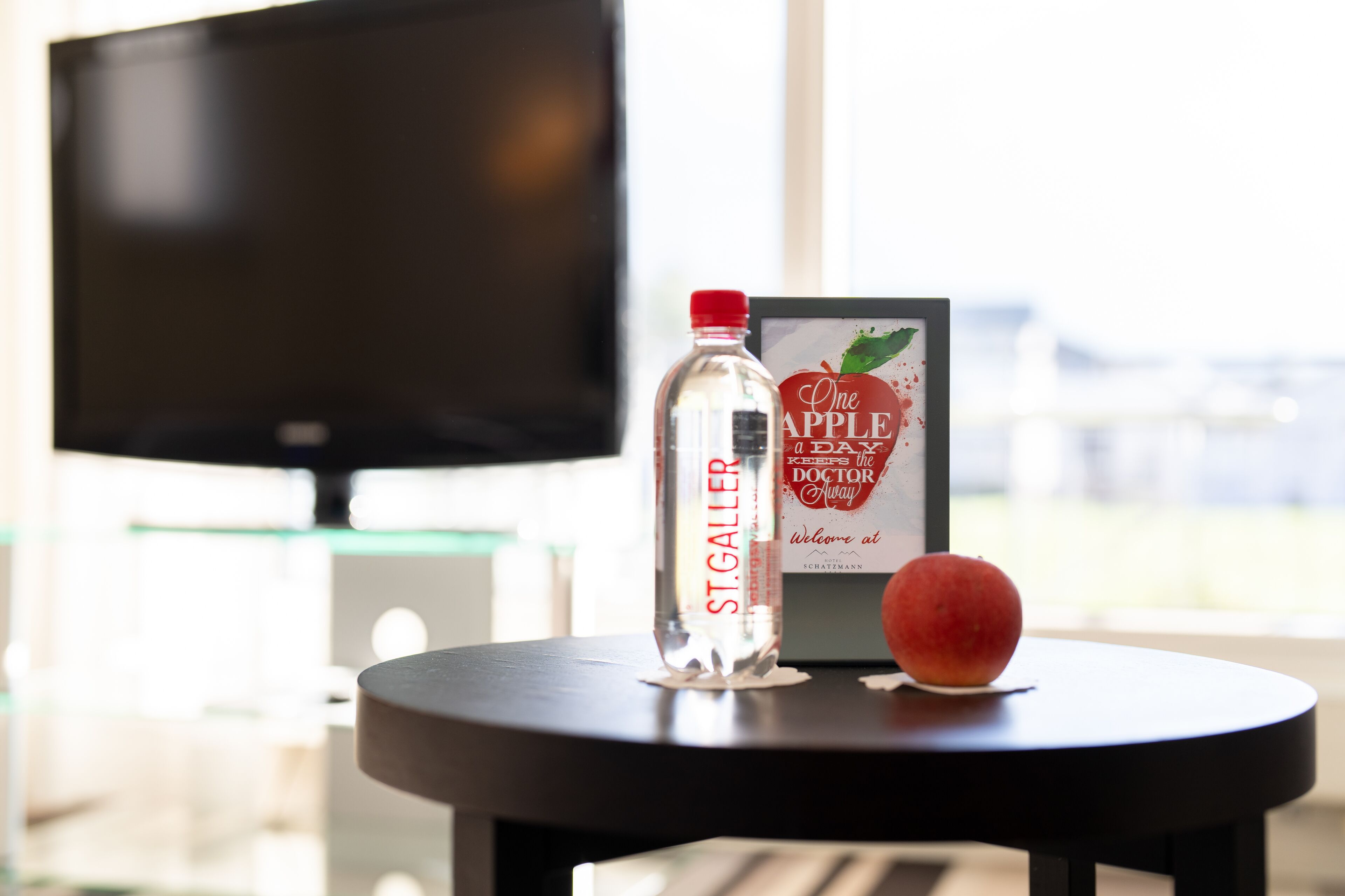 A bottle of St. Galler water, a framed sign, and a fruit on a table in a room with a TV in the background.