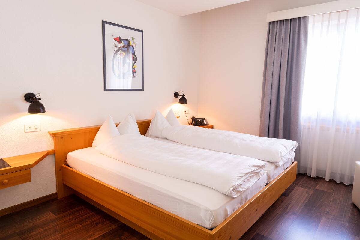 Ein Doppelbett mit weißen Leinentüchern in einem Zimmer mit Holzboden, zwei Nachttische mit Lampen, ein gerahmtes Bild an der Wand und ein Fenster mit Vorhängen.