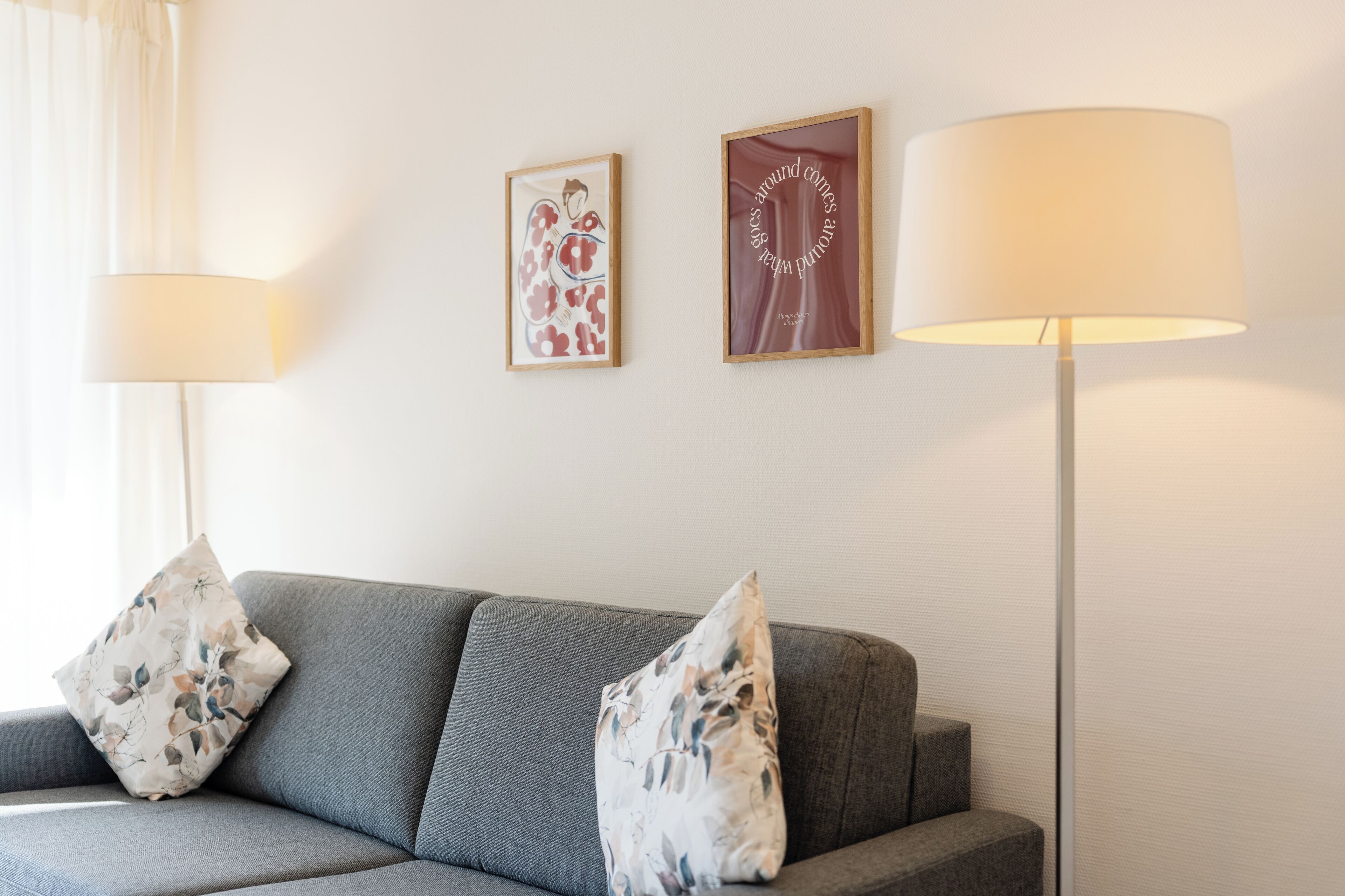 Un sofà gris amb coixins de motius florals, una làmpada de peu i dos quadres emmarcades a la paret.