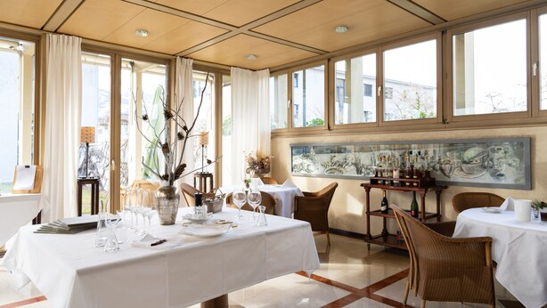 A dining area with a table set for four, a large window with curtains, and a painting on the wall. - Hotel Schatzmann (Triesen)