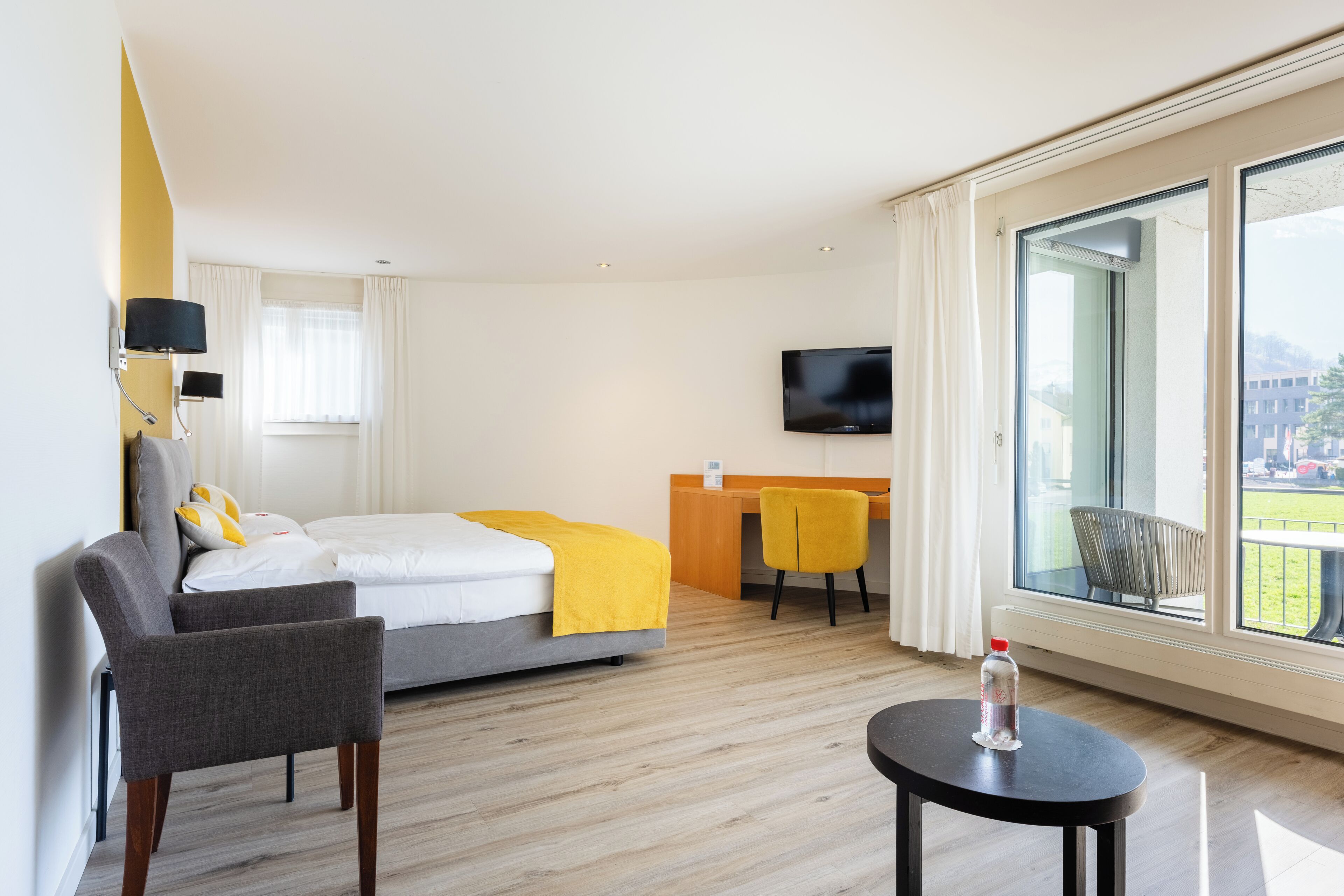 Hotellihuone, jossa on sänky, työpöytä, tuoli, televisio ja suuri ikkuna.