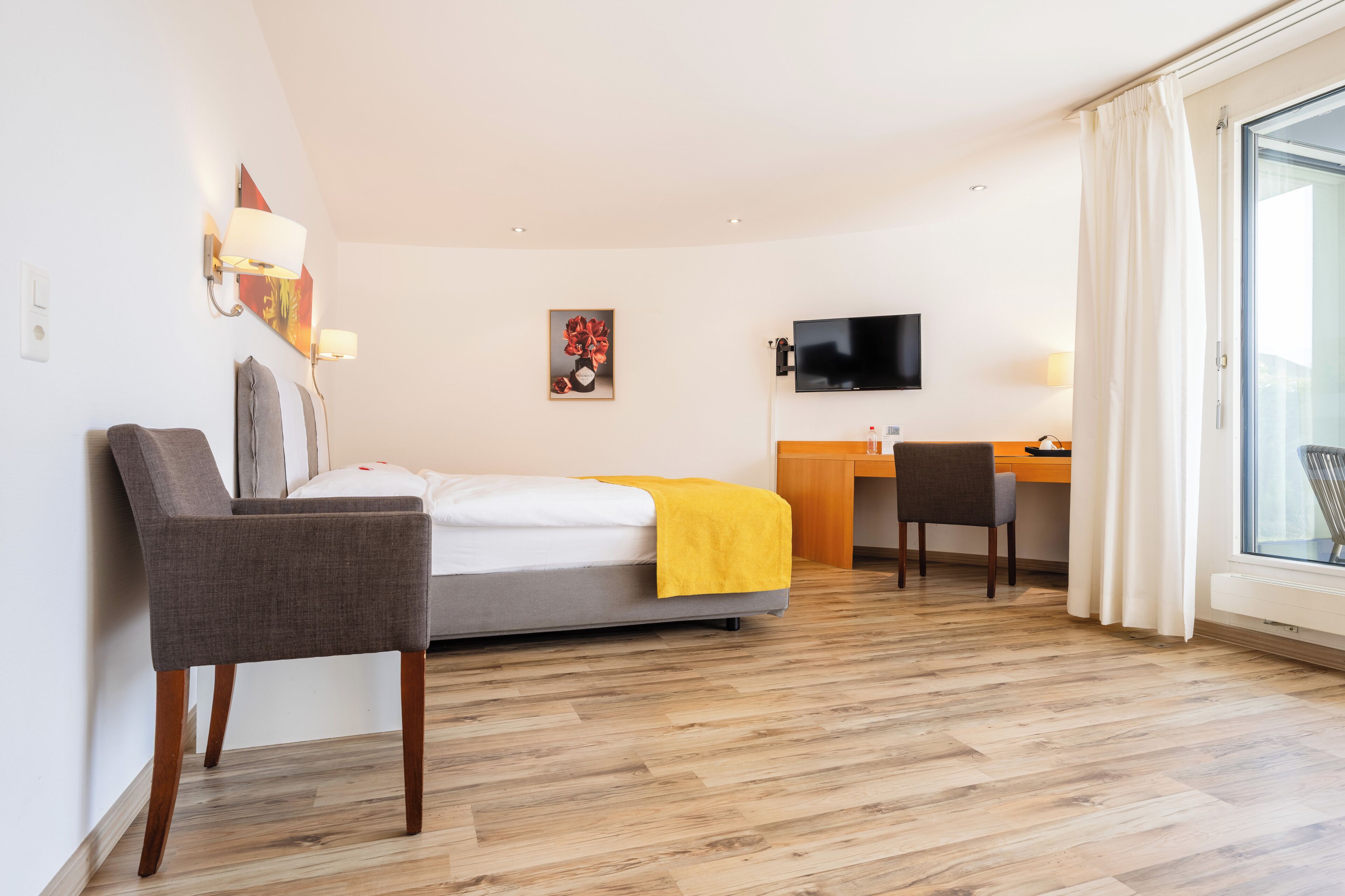 A hotel room with a bed, a desk, a chair, a TV, and a large window with curtains.