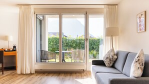 A room with a grey sofa, a desk with a lamp, and a large sliding glass door leading to a balcony with a view of greenery and mountains. - Hotel Schatzmann (Triesen)