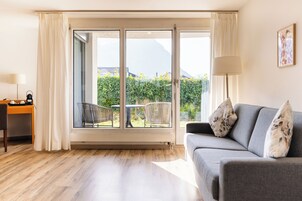 Una habitación con un sofá gris, un escritorio con una lámpara y un amplio ventanal corredera que da a un balcón con vistas a zonas verdes y montañas.