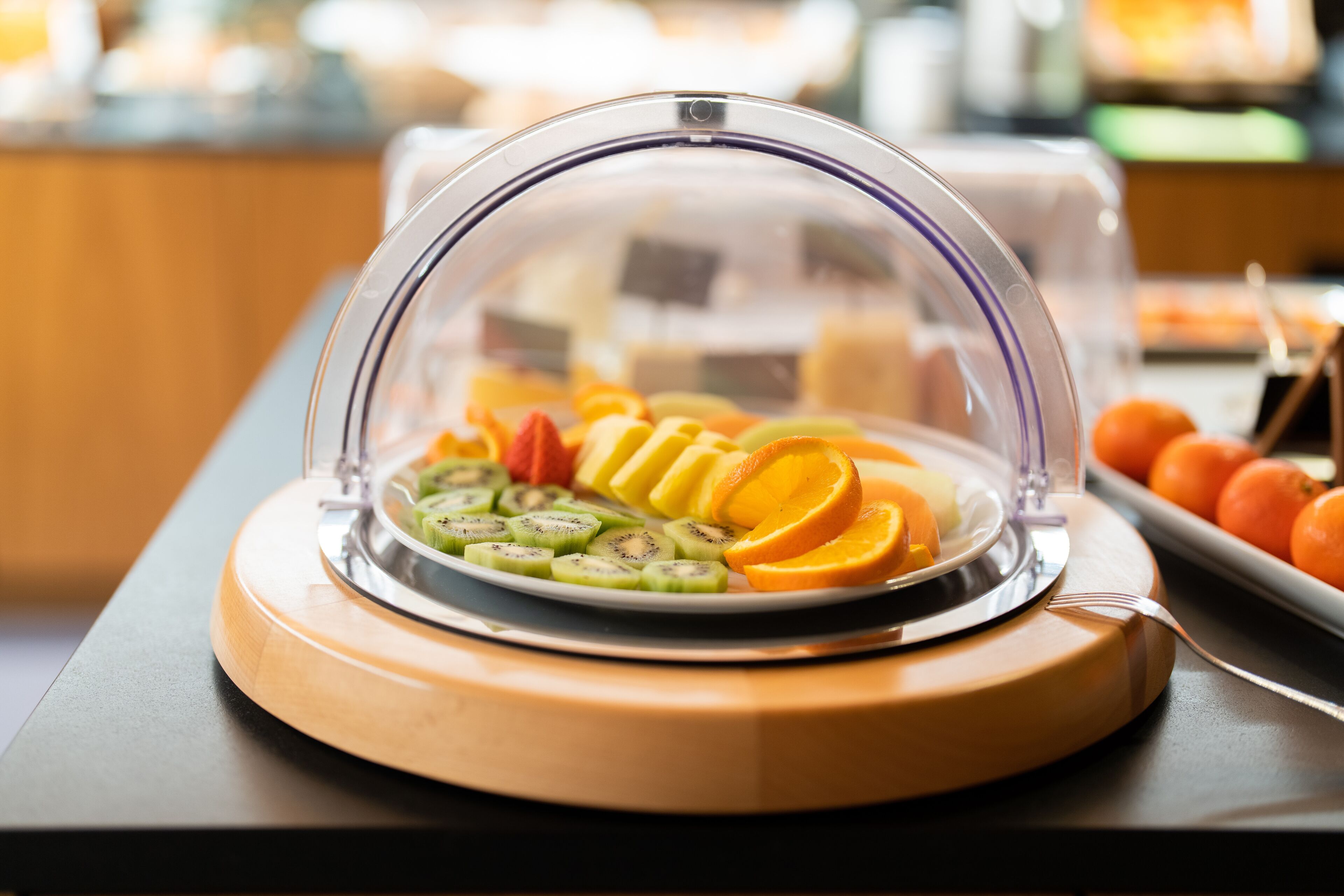 Un bufé con una variedad de frutas cortadas, incluyendo kiwi, naranja y melón, cubierto por una cúpula transparente.
