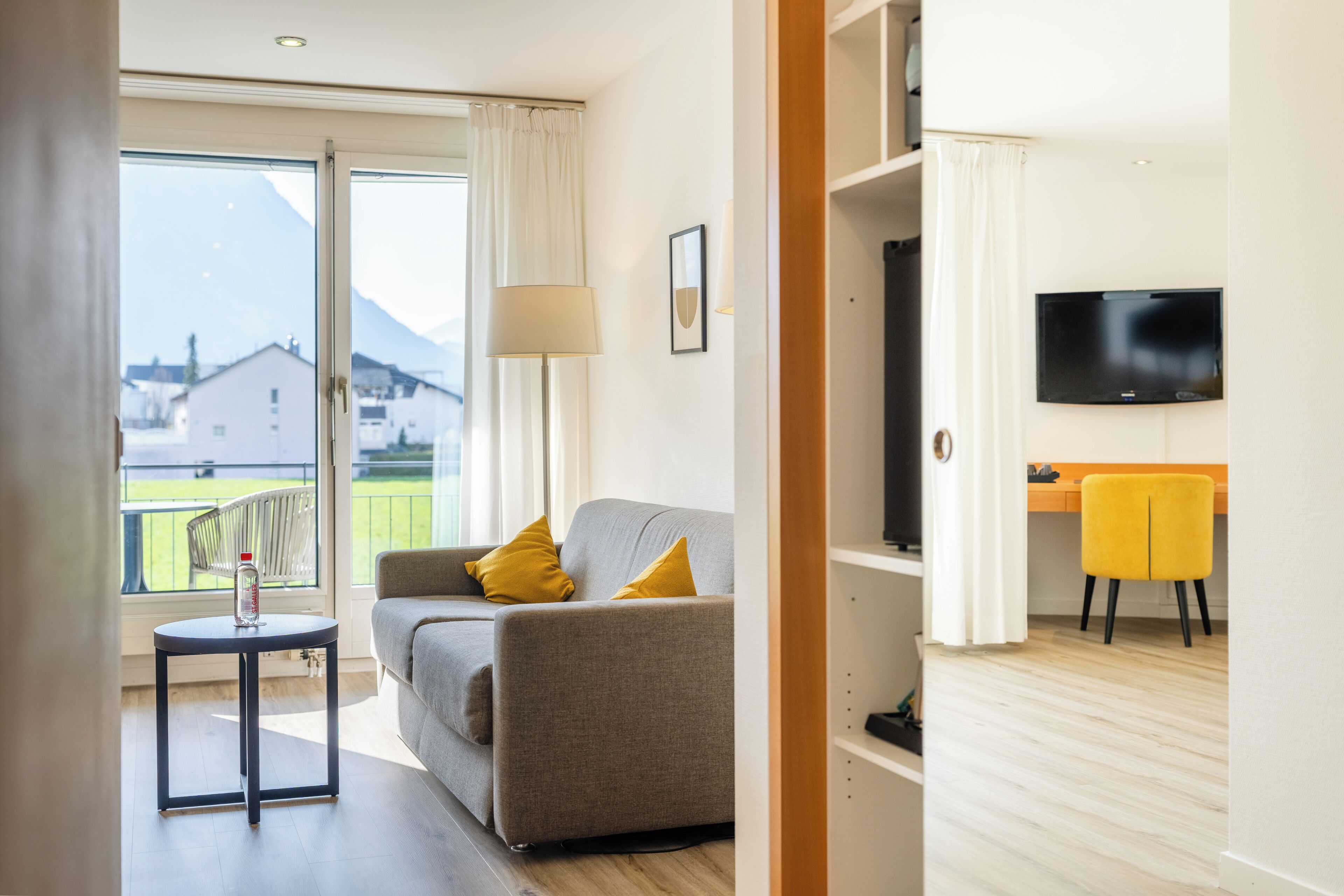 A modern living room with a sofa, a small table, a TV, and a balcony with a view of houses and mountains.