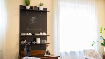 A cozy hotel room with a wooden desk, a chalkboard with a coffee-related message, a small shelf with plants, and a window with sheer curtains.