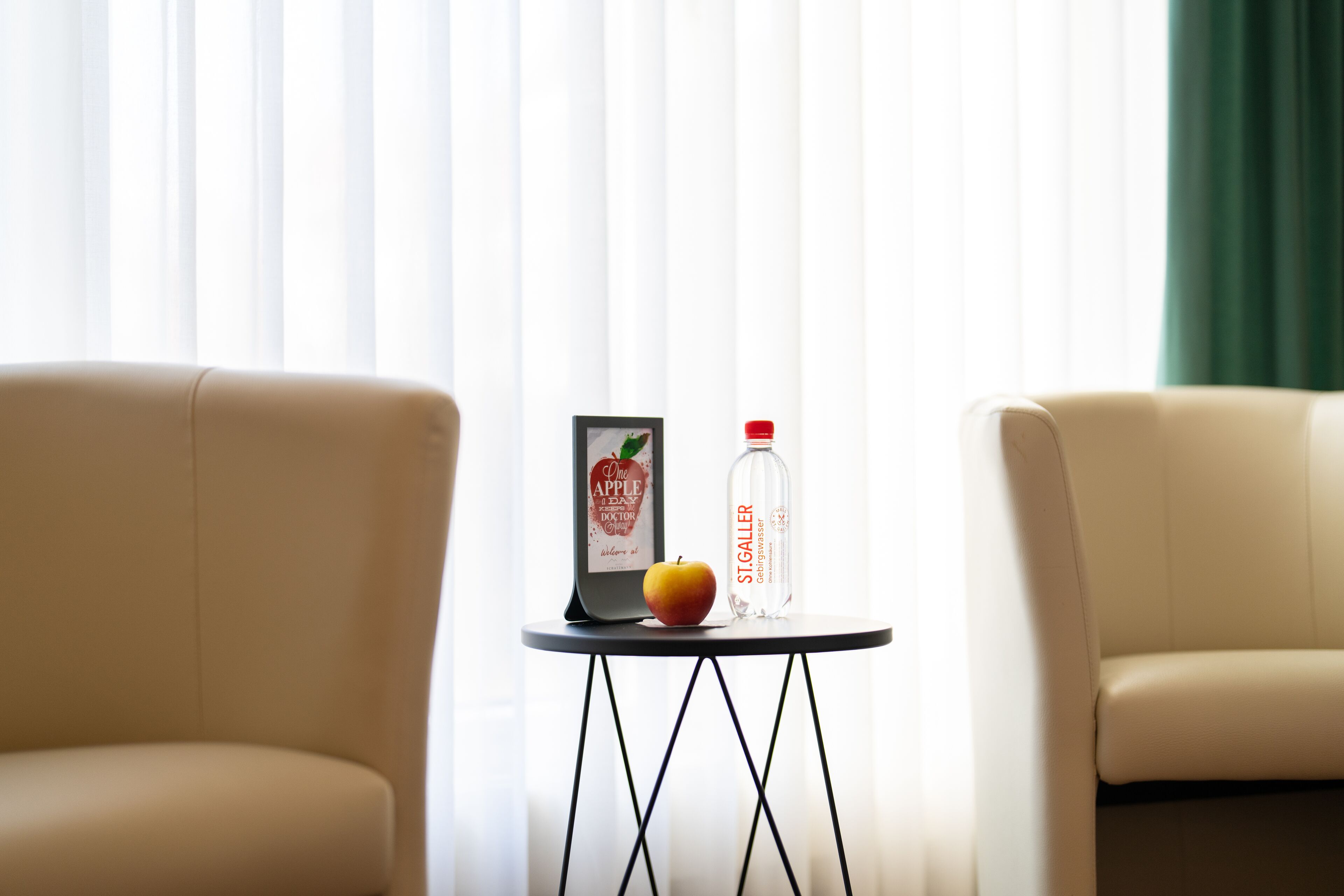 A room with two beige armchairs, a small table with a framed picture, a bottle of St. Galler, and an apple.