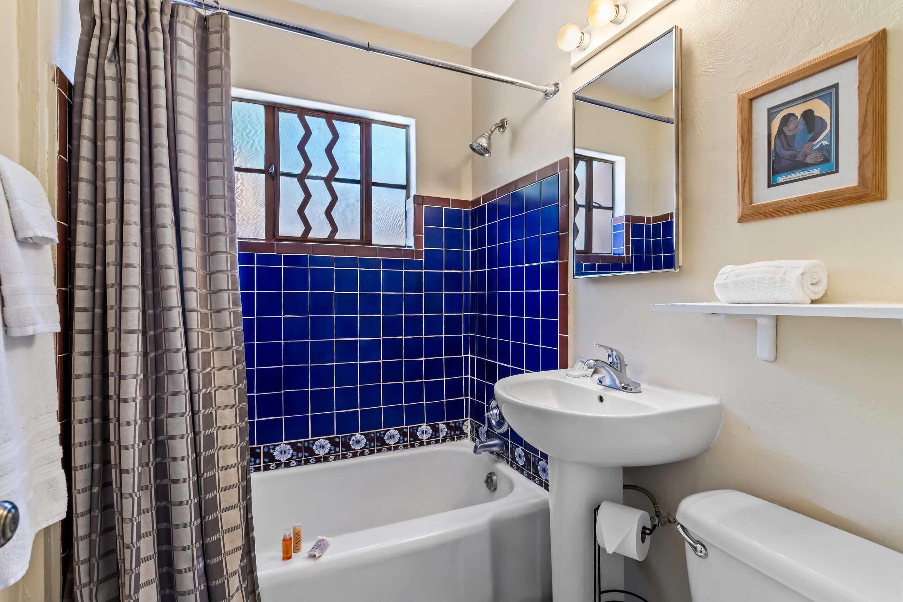 Un baño con bañera, lavamanos e inodoro. Hay una cortina de ducha, una ventana con un diseño en zigzag y un cuadro en la pared.