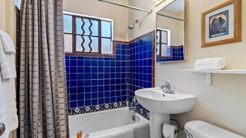 Un baño con bañera, lavamanos e inodoro. Hay una cortina de ducha, una ventana con un diseño en zigzag y un cuadro en la pared.