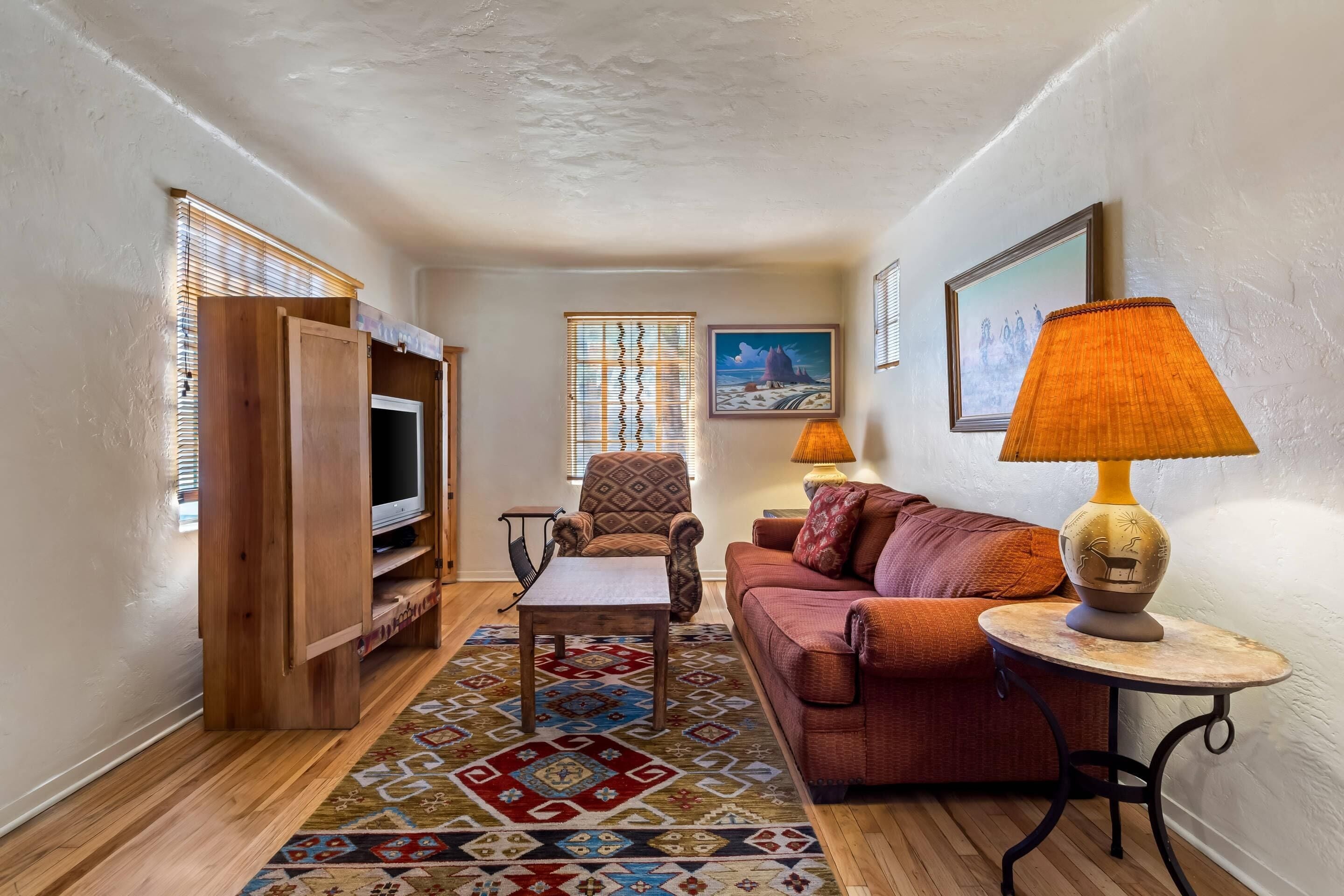 A cozy living room with a wooden entertainment unit, a patterned rug, a red sofa, a small round table with a lamp, and a framed painting on the wall.