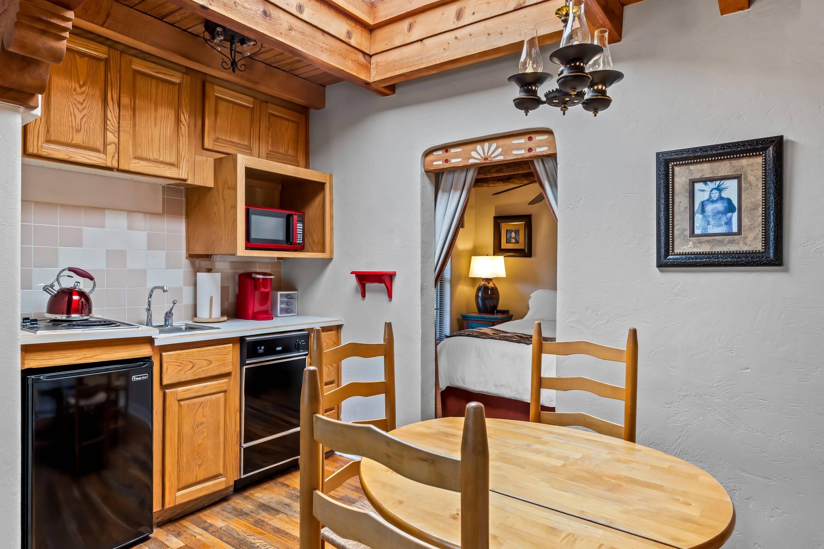 A compact kitchen with wooden cabinets, a microwave, a red kettle, and a dining area with a wooden table and chairs.