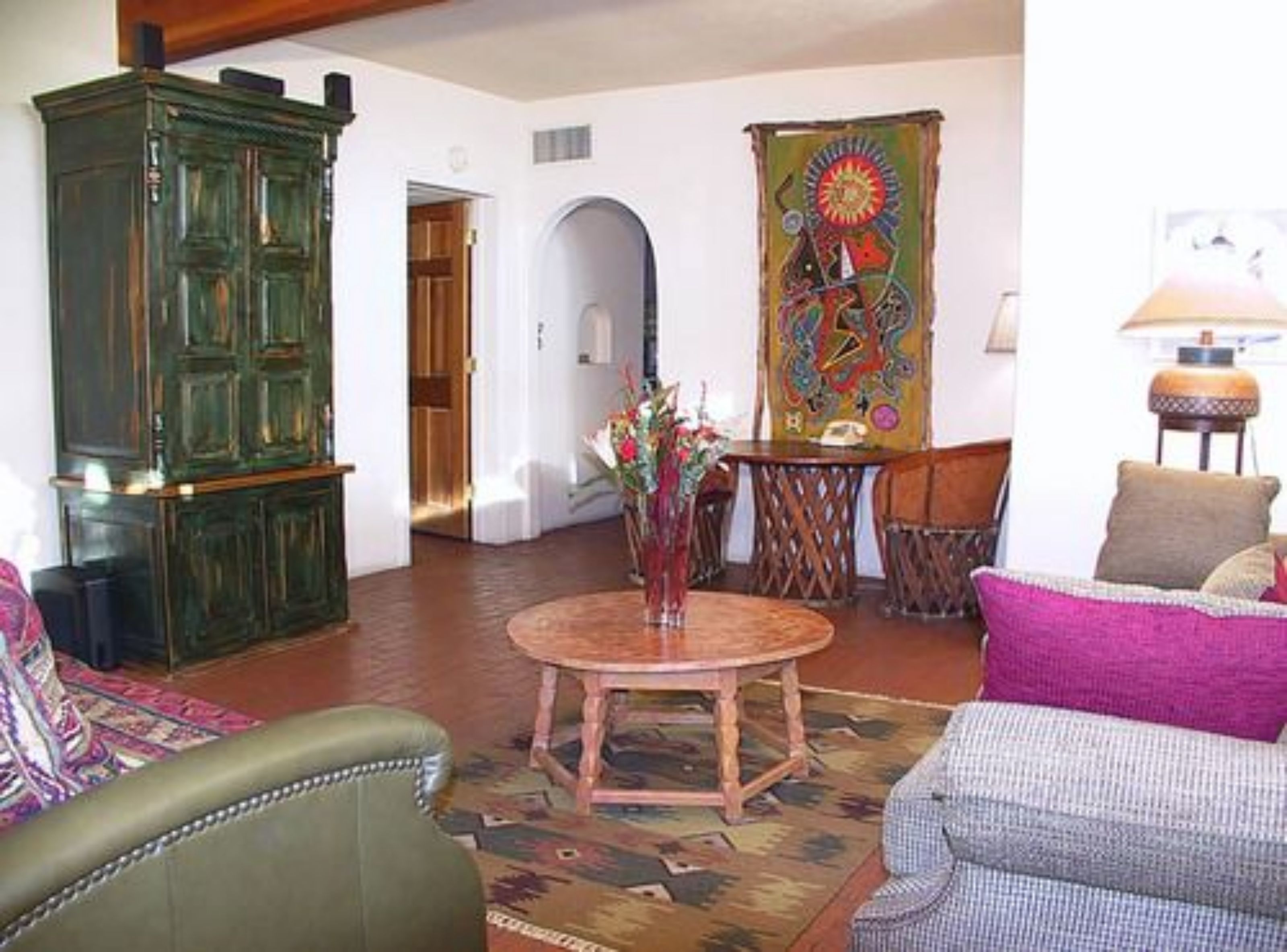 A living room with a green cabinet, a round wooden table, a colorful tapestry, and a lamp.