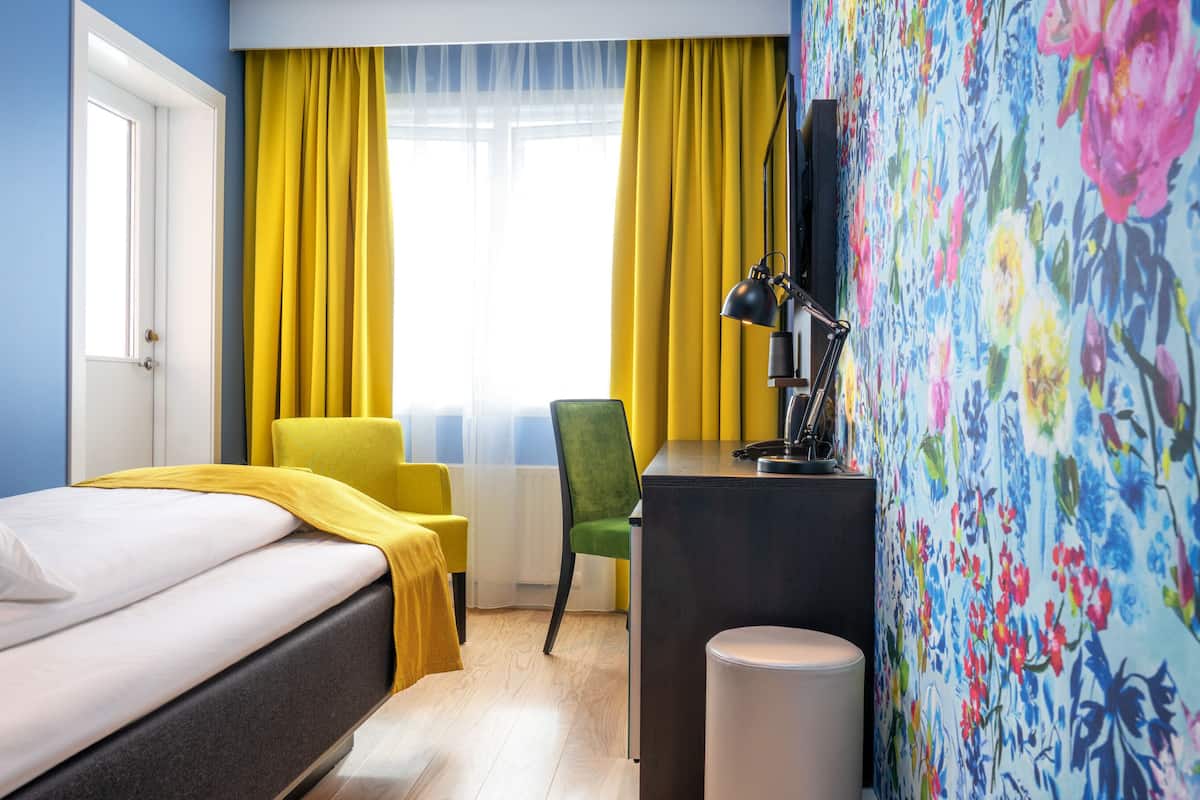 A hotel room with a bed, a desk, a chair, and a window with yellow curtains.
