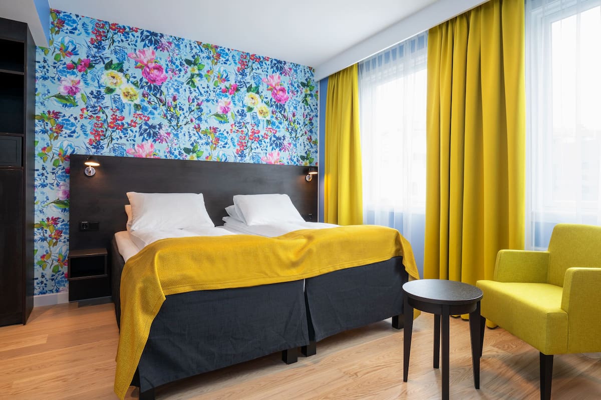 A hotel room with a bed, a small table, a yellow armchair, and a floral wallpaper.