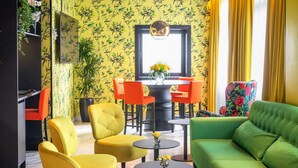Une pièce avec un papier peint jaune à motifs, une table à manger entourée de chaises orange, un canapé vert et une petite table ronde sur laquelle est posé un vase de fleurs.