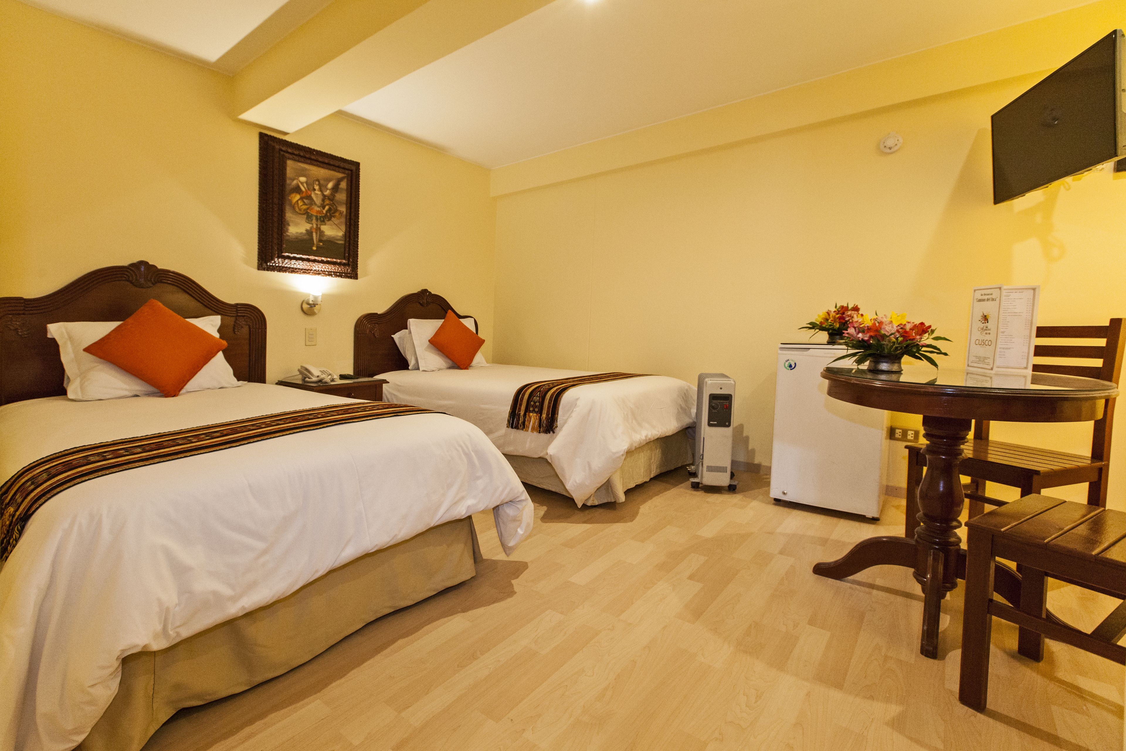 A hotel room with two beds, a small table with a flower vase, a refrigerator, and a TV mounted on the wall.