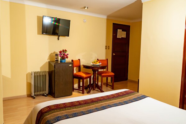 Une chambre d’hôtel avec un lit, une petite table avec des chaises, un radiateur, un téléviseur fixé au mur et une porte sur laquelle est affiché un avis.