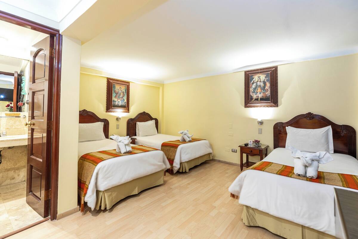 A hotel room with two beds, a wooden door, a small table with a lamp, and a painting on the wall.