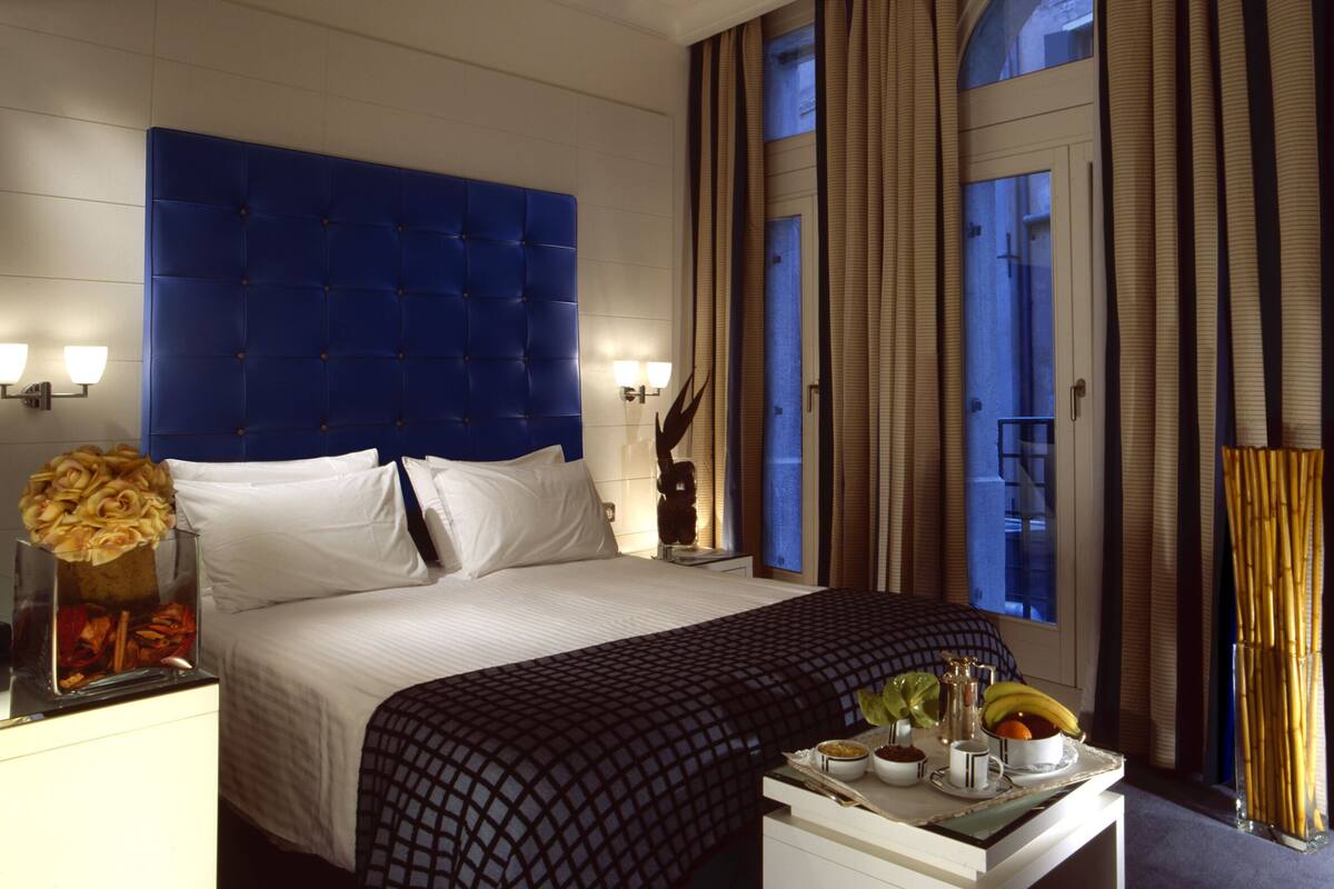 A hotel room with a large bed, a nightstand with a vase of flowers, a tray with fruits and a coffee pot, and a window with curtains.