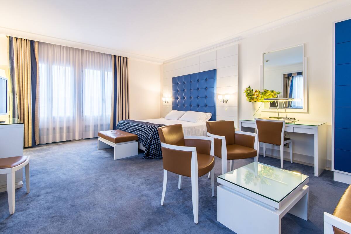 A modern hotel room with a bed, two chairs, a desk, and a glass table.