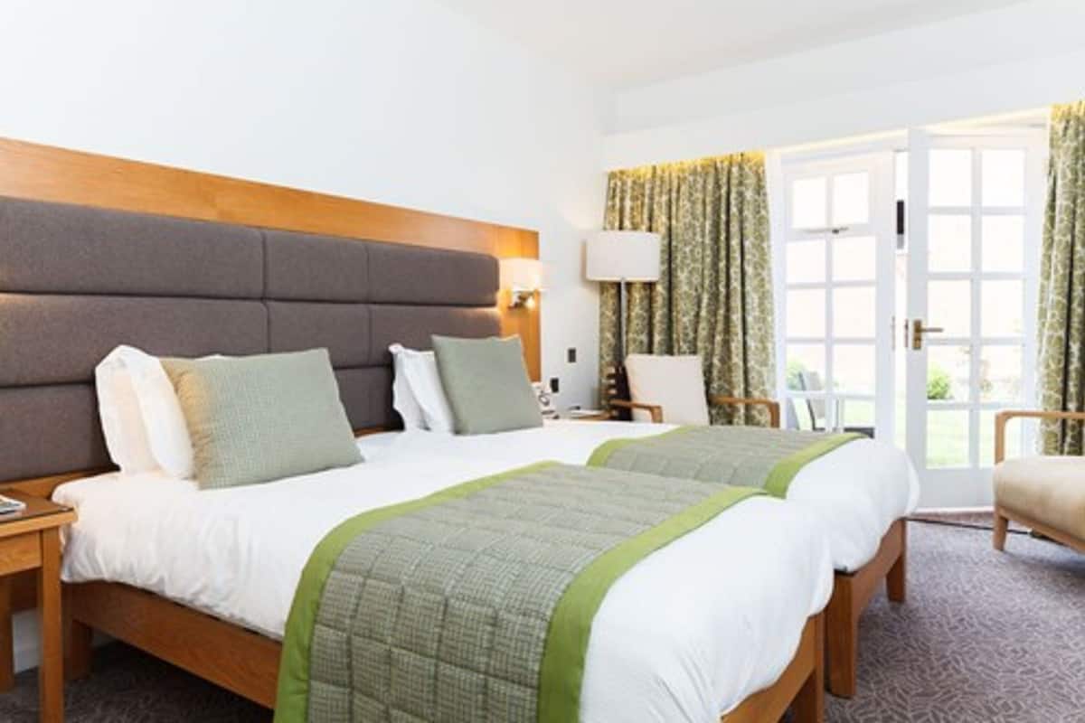 A hotel room with two beds, a wooden headboard, a nightstand, a lamp, a chair, and a view of the outdoors through a door.