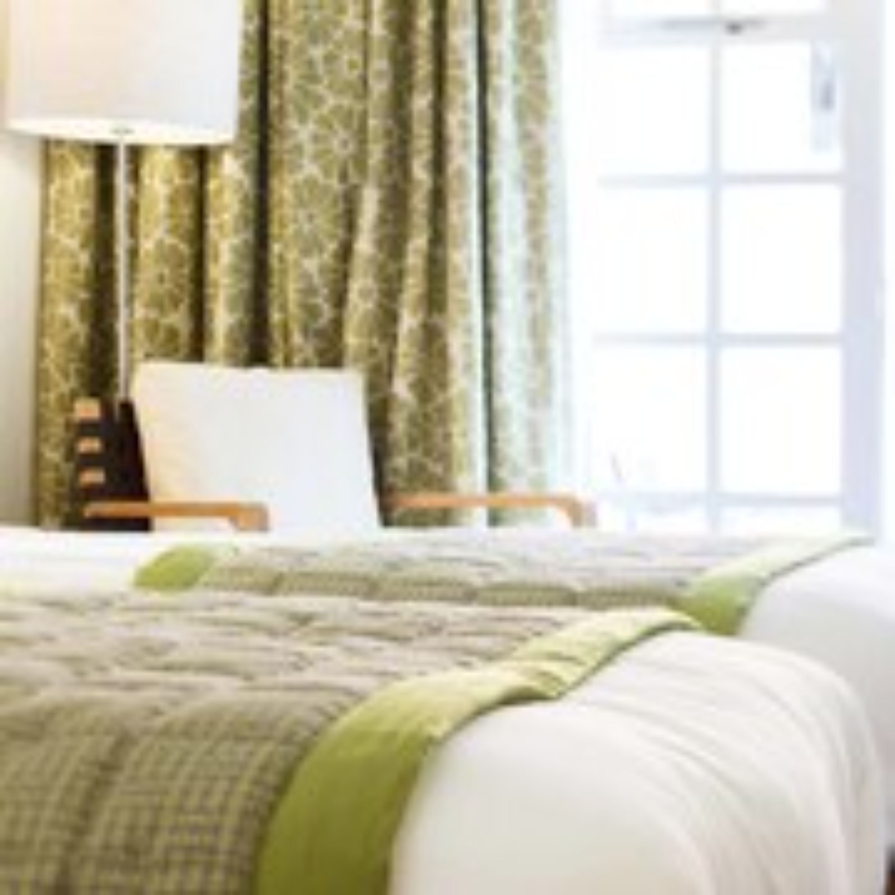 Quarto de hotel com cama, mesa de cabeceira, abajur e janela com cortinas.