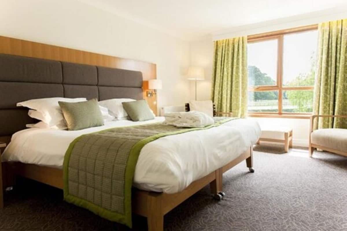 A hotel room with a large bed, a wooden headboard, a bedside table with a lamp, a chair, and a window with curtains.