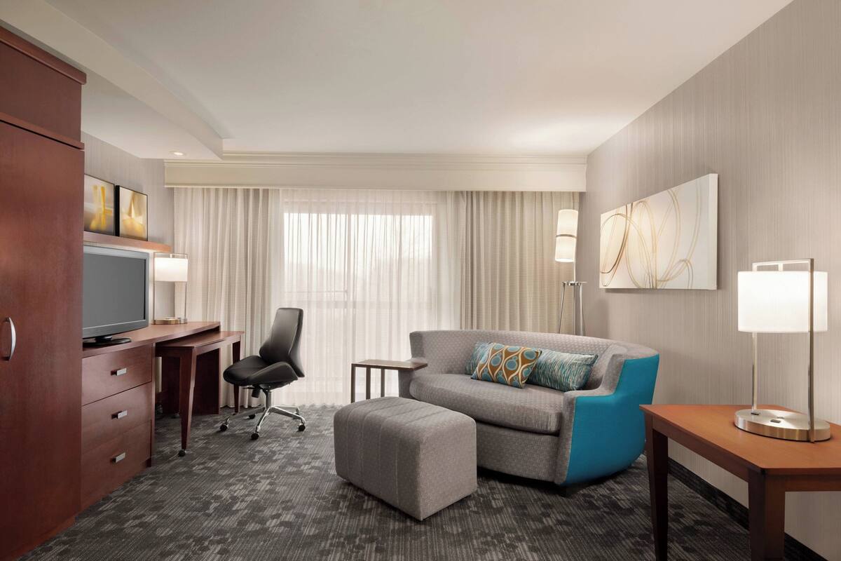 Une chambre d’hôtel équipée d’un bureau, d’une télévision, d’un canapé, d’un pouf, d’une lampe et d’un tableau au mur.