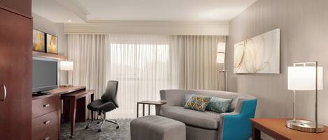 A hotel room with a desk, a television, a sofa, an ottoman, a lamp, and a painting on the wall.