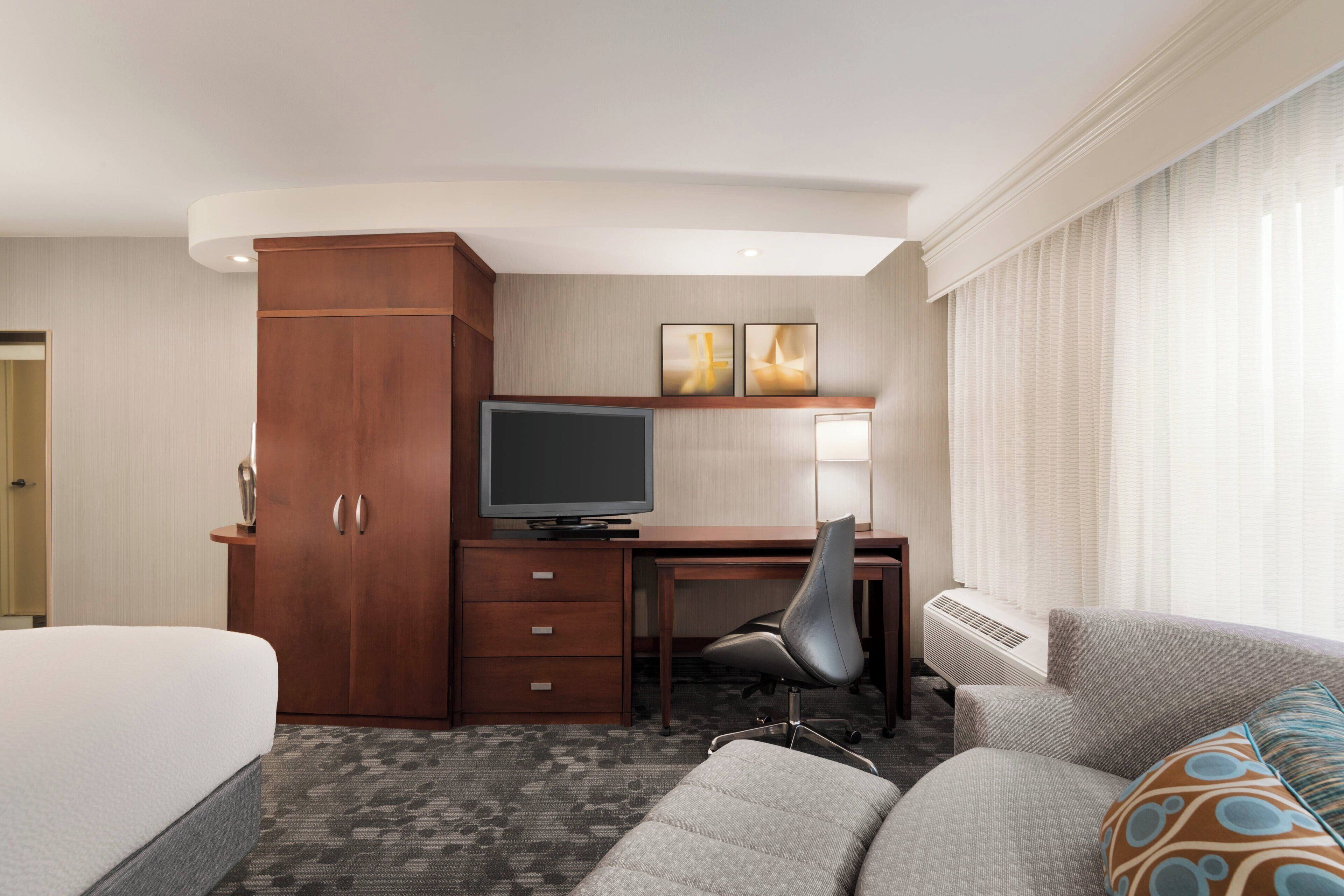 Quarto de hotel com cama, escrivaninha com computador, cadeira, sofá e janela com cortinas.