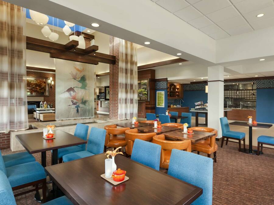 A hotel lobby with multiple seating areas, including a central dining table surrounded by blue chairs and a smaller table with orange chairs. There are decorative elements like a vase and a candle on the central table. The lobby features a large mural on the wall and a brick accent wall.