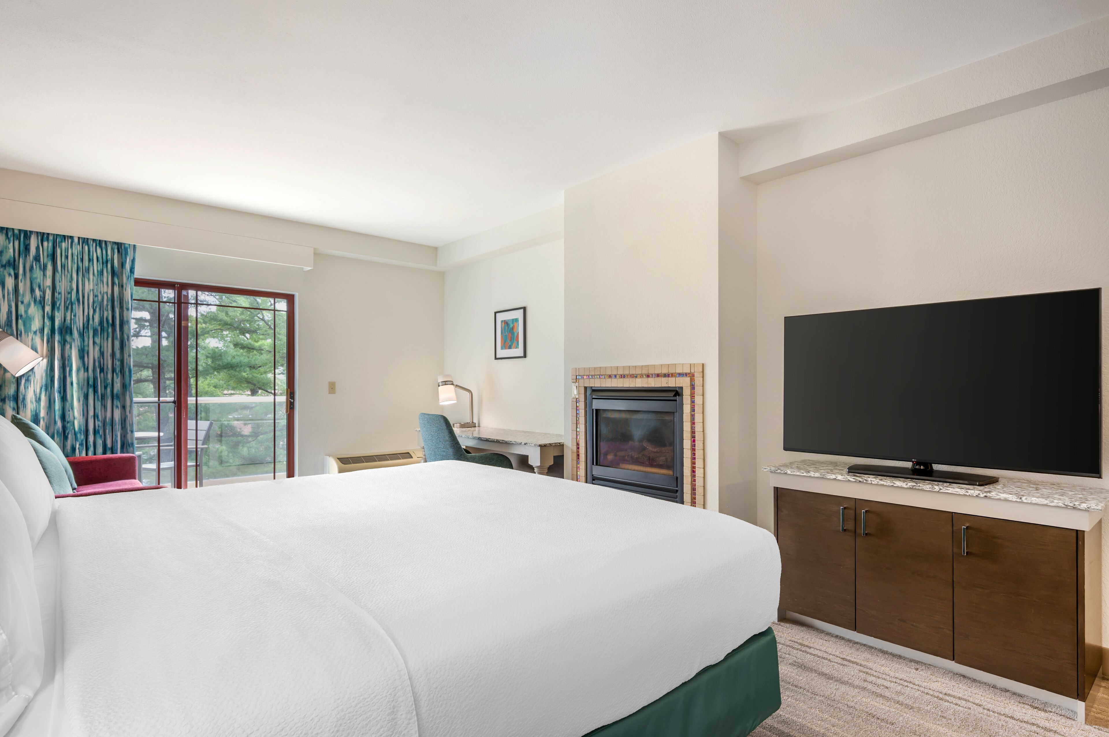 A hotel room with a large bed, a flat-screen TV, a fireplace, and a balcony with curtains.