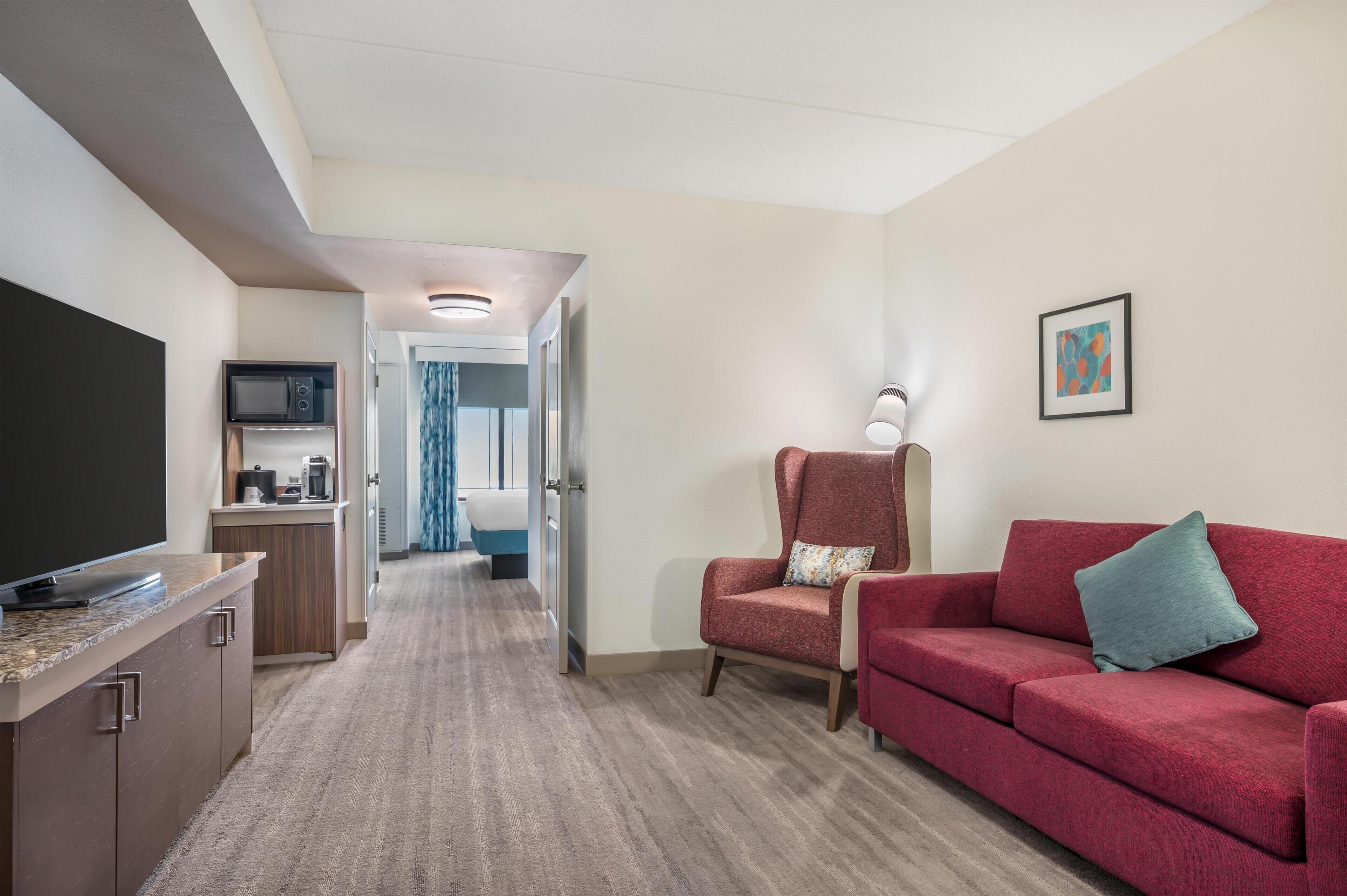 A hotel room with a kitchenette, a red sofa, a red armchair, a flat-screen TV, and a framed picture on the wall.