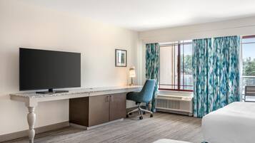 Une chambre d’hôtel équipée d’une grande télévision à écran plat, d’un bureau avec une chaise, d’un lit, d’une fenêtre avec des rideaux et d’un tableau encadré au mur.