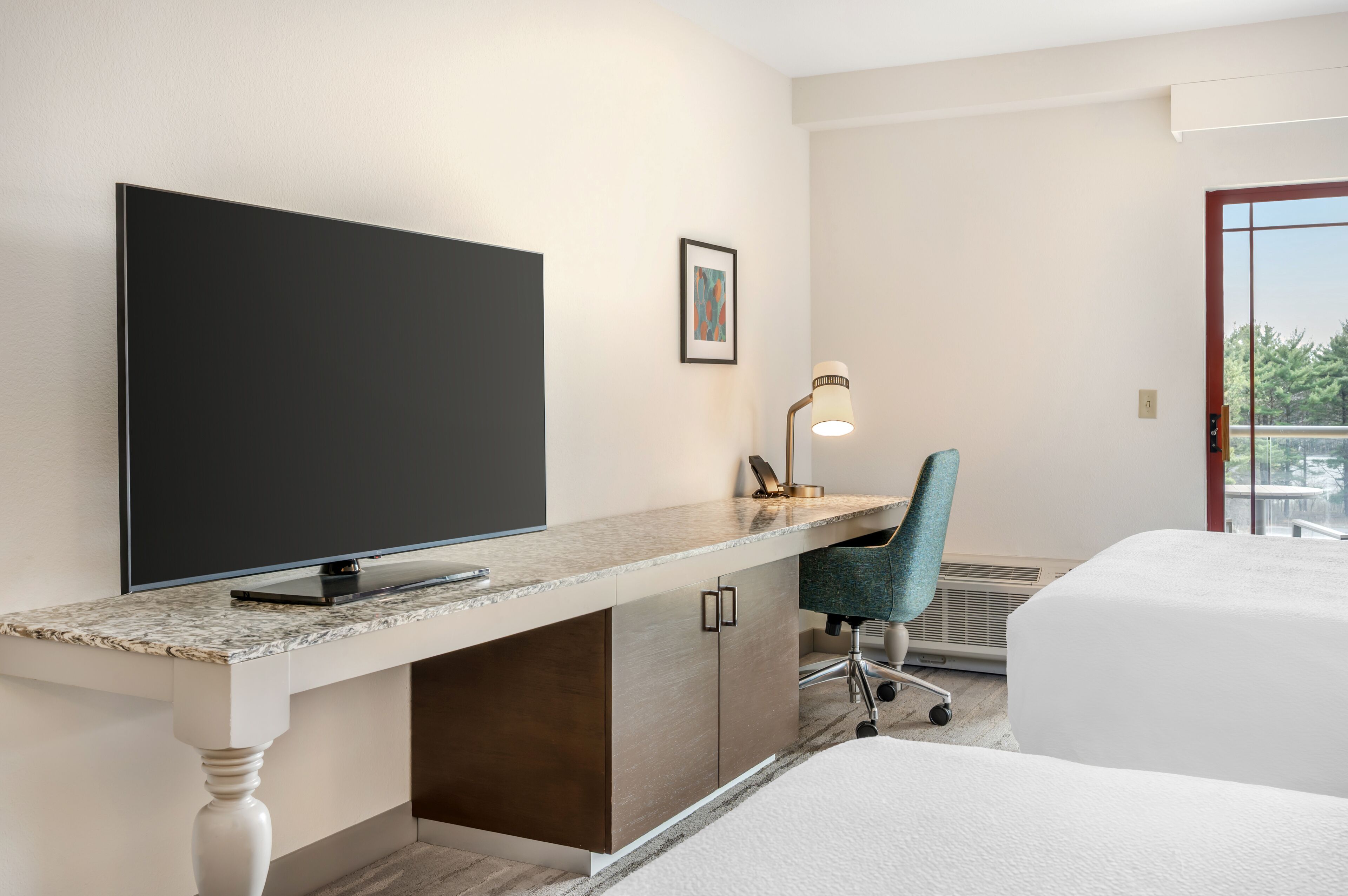 A hotel room with a large flat-screen TV, a desk with a lamp, a chair, and a bed.