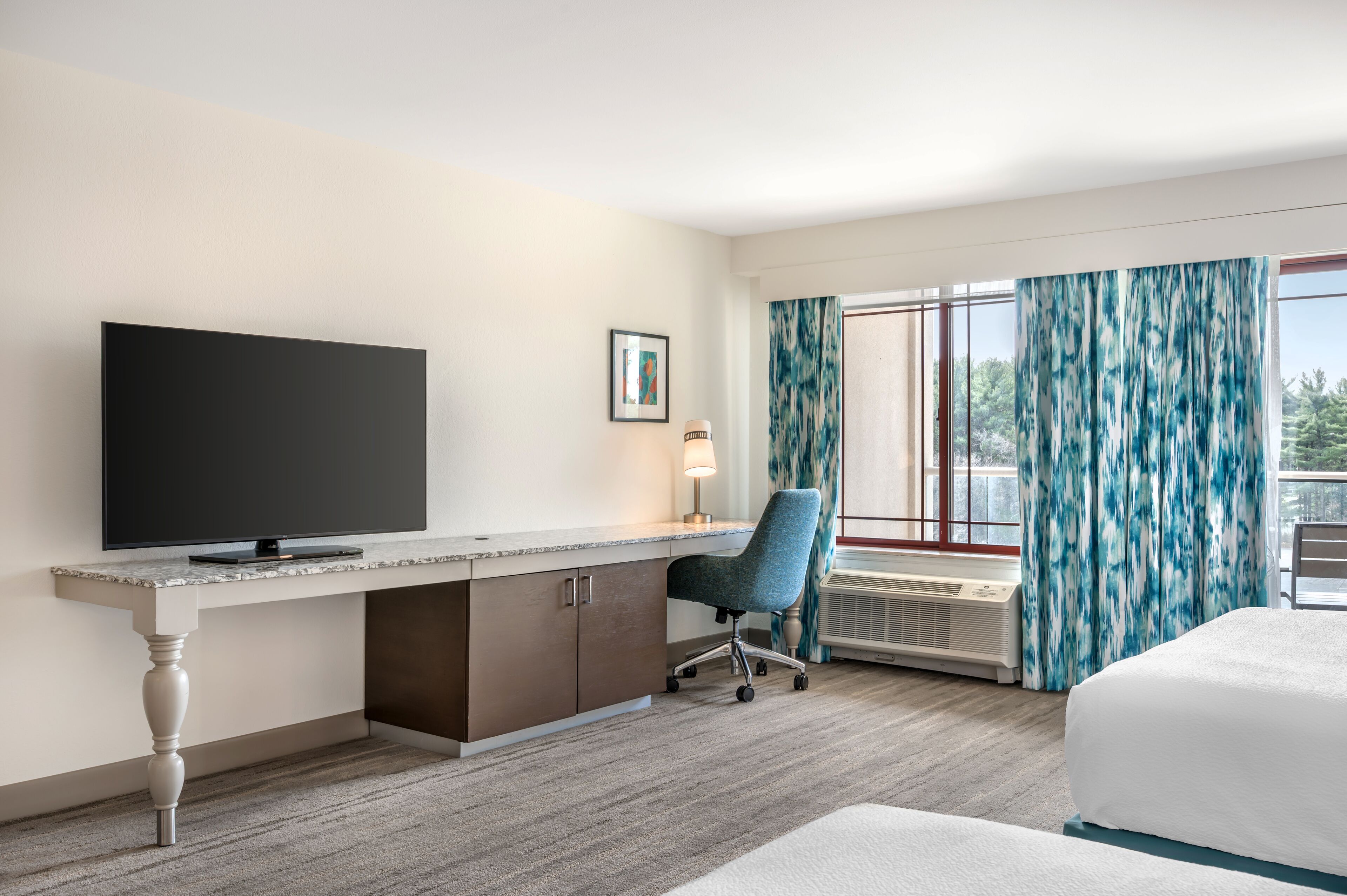 Quarto de hotel com televisão de tela plana, escrivaninha com cadeira, cama e uma grande janela com cortinas.