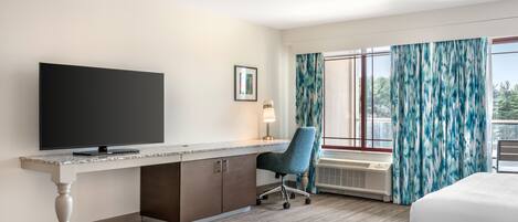 Et hotelværelse med et fladskærms-tv, et skrivebord med stol, en seng og et stort vindue med gardiner.