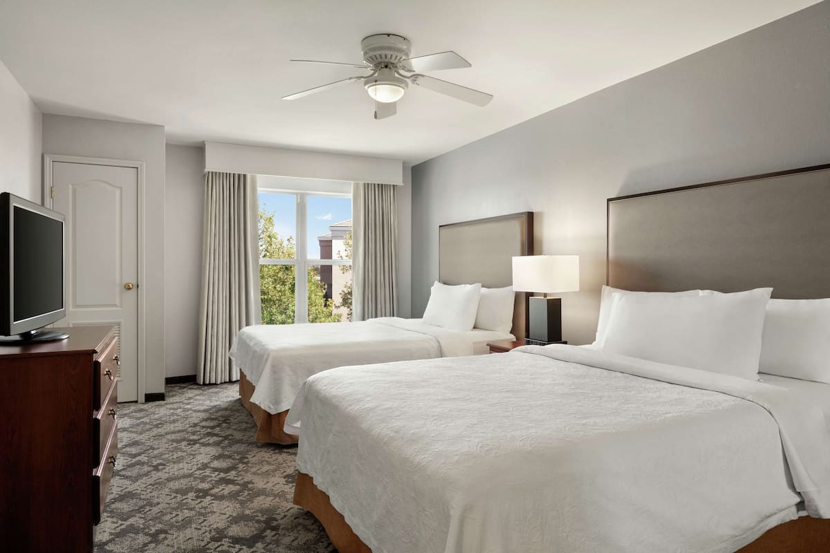 A hotel room with two beds, a ceiling fan, a TV, and a view of the outdoors.