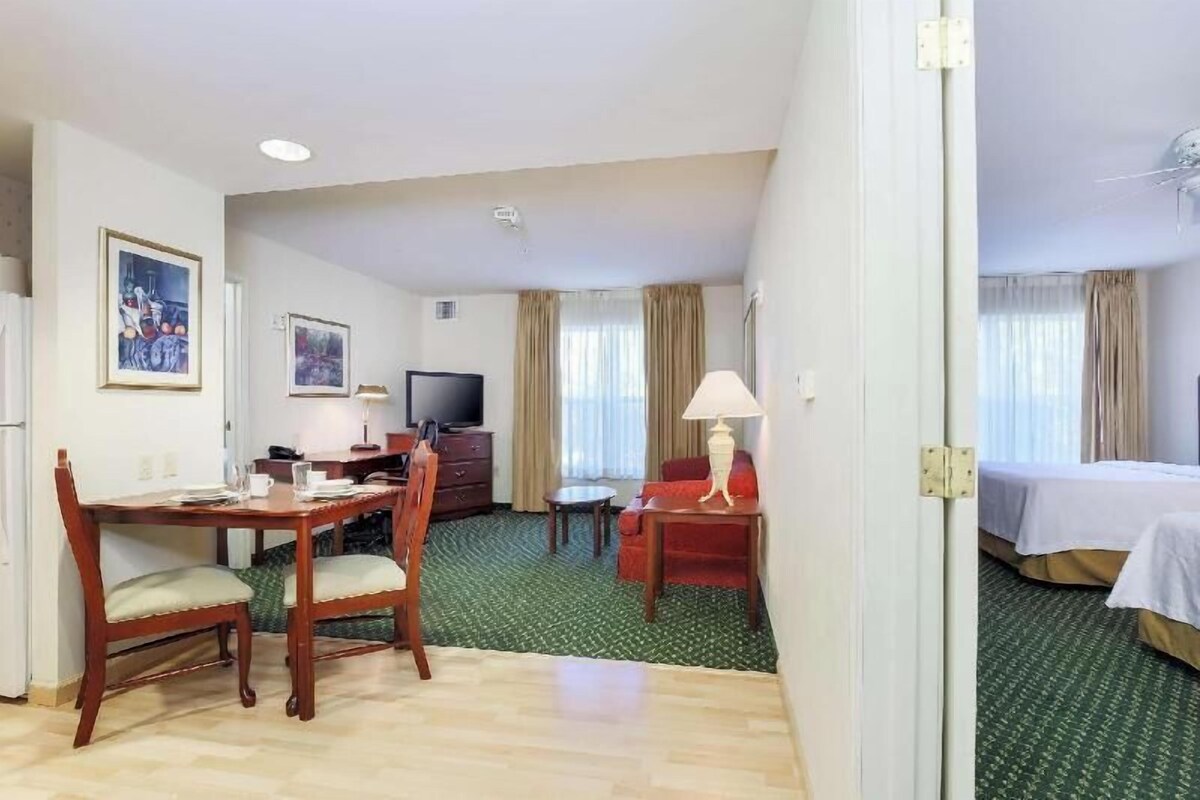 Habitación de hotel con una mesa de comedor, sillas, televisor, cama y una mesita de noche con lámpara.