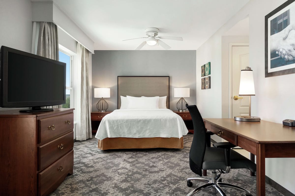 A hotel room with a bed, two bedside lamps, a desk with a chair, a television on a dresser, and a ceiling fan.