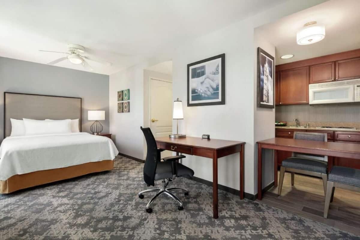 A hotel room with a bed, desk, chair, kitchenette, and ceiling fan.