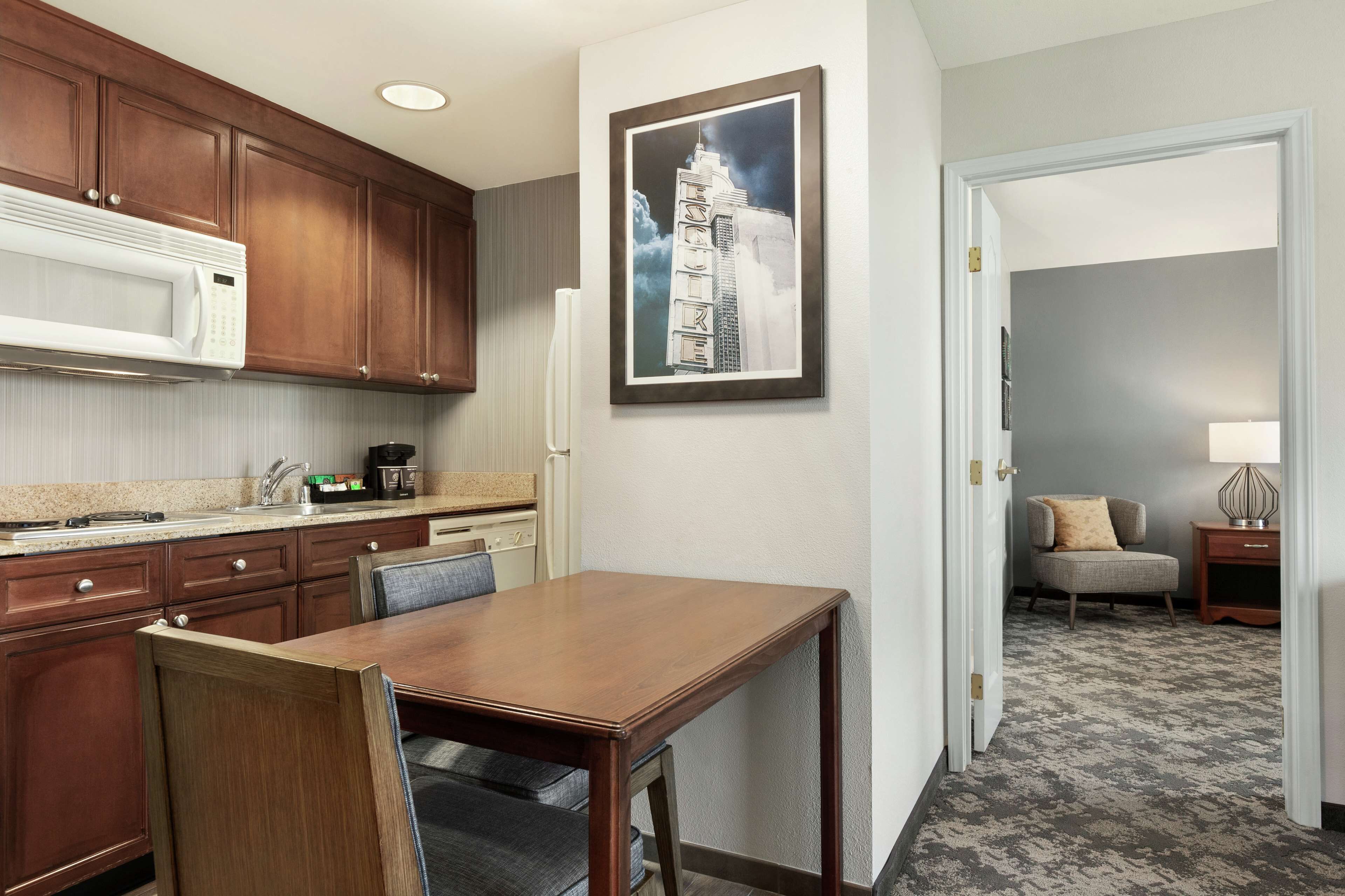 A hotel room with a dining area, a microwave, a framed picture on the wall, a chair, a lamp, and a carpeted floor.