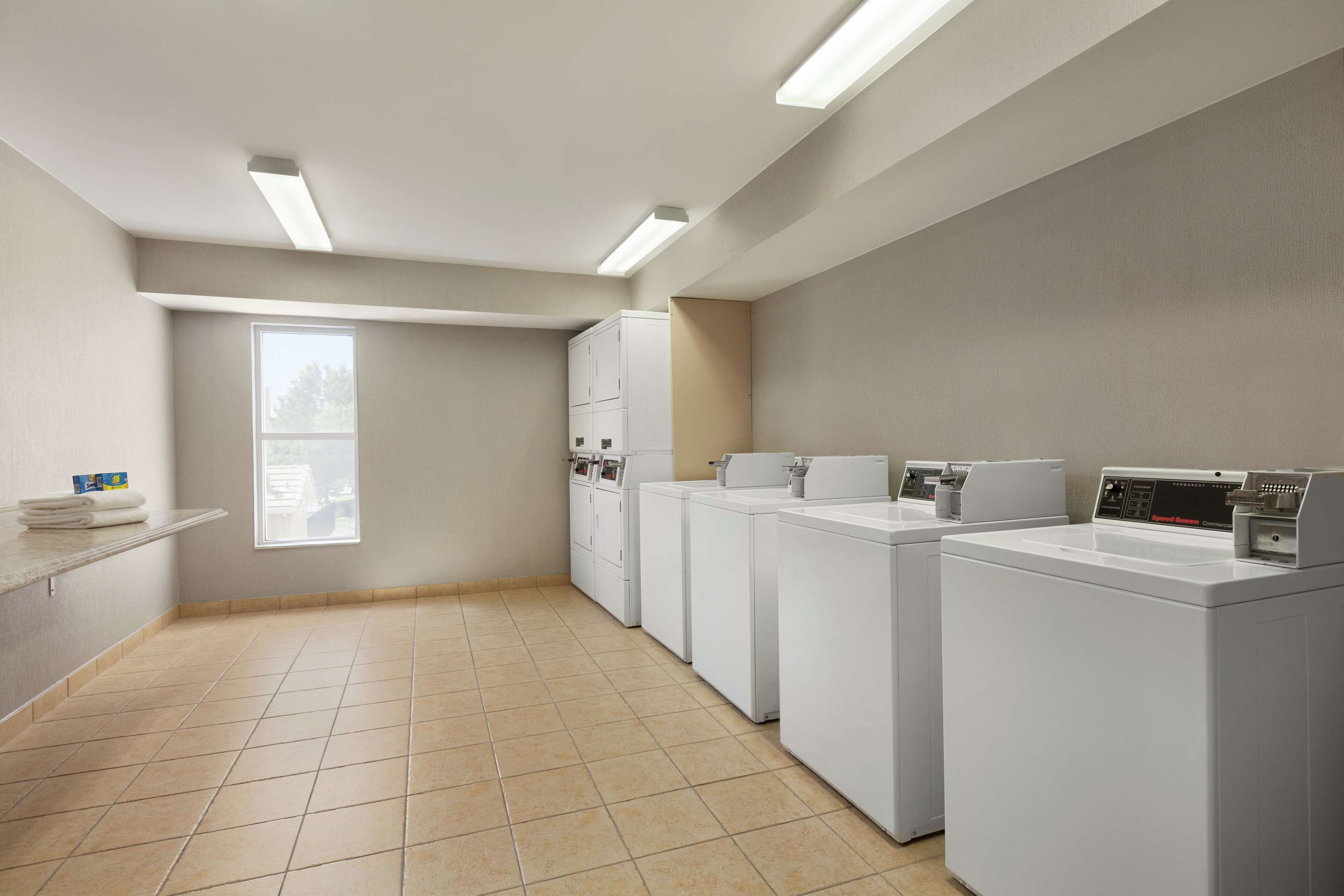 A laundry room with four white washing machines, a large window, and a tiled floor.