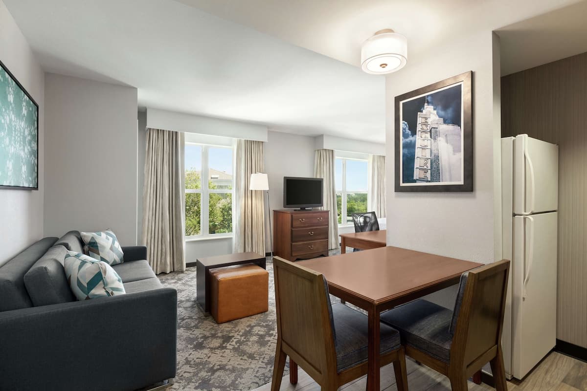 A hotel room with a sofa, a wooden dining table with chairs, a television, a refrigerator, and a large window with curtains.