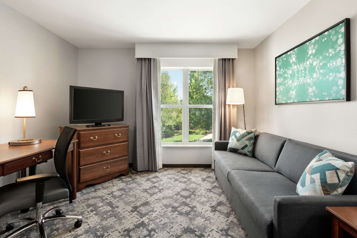A hotel room with a flat-screen TV on a wooden dresser, a grey sofa with patterned cushions, a desk with a lamp, and a large window with curtains.