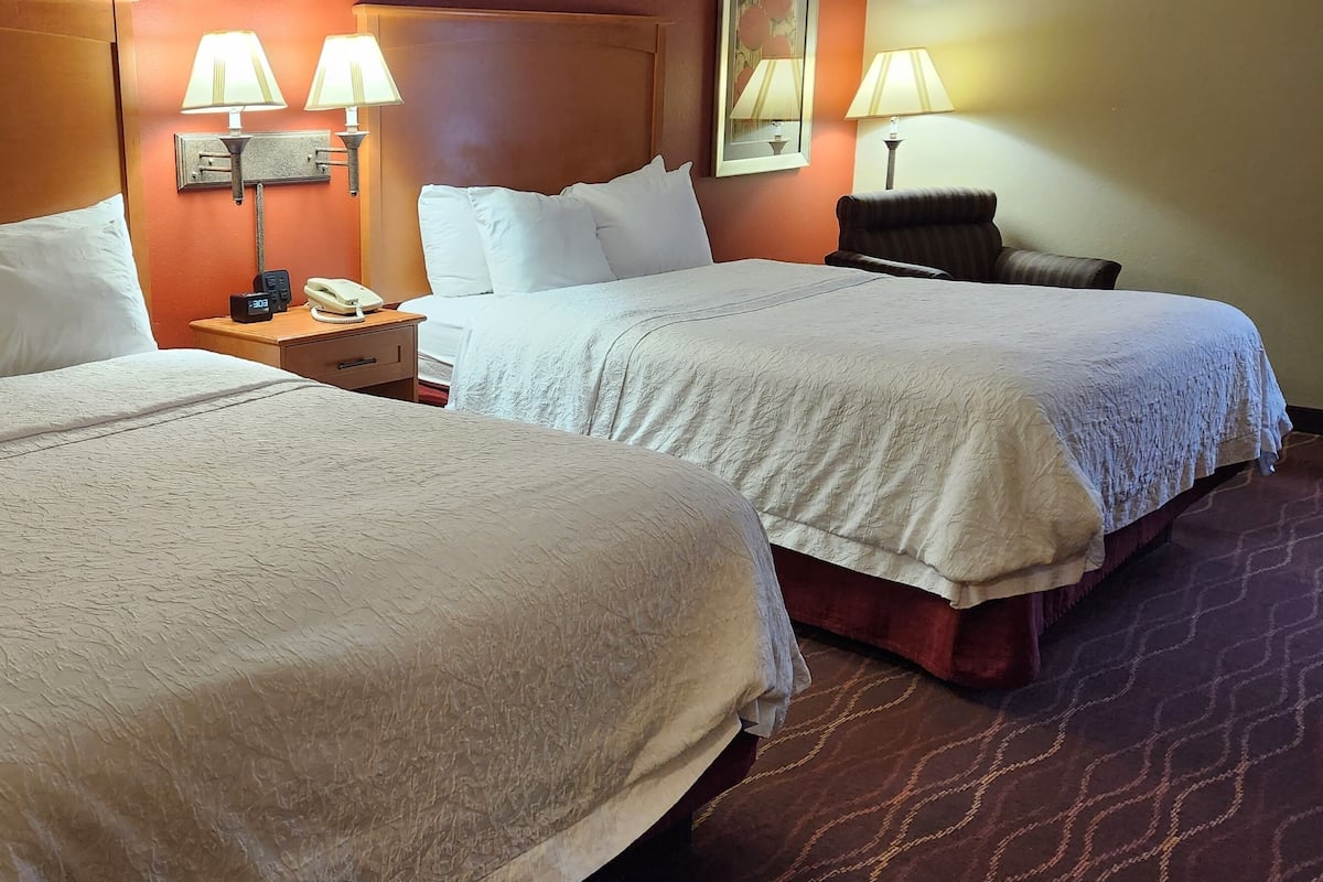 A hotel room with two beds, a nightstand with a phone and lamp, and a chair.